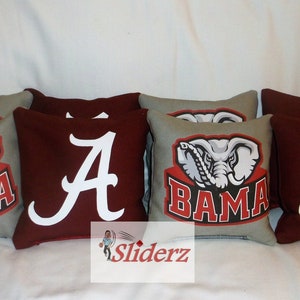 May include: Set of eight bean bags for a game of cornhole. The bean bags are made of fabric and have a red and gray color scheme. Four bean bags have a white elephant logo with the word "BAMA" underneath. The other four bean bags have a white letter "A" on a red background.