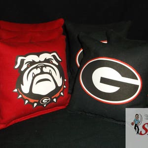 May include: A set of cornhole bags in red and black. The red bags feature a white and black bulldog graphic. The black bags have a white and red "G" logo. The bags are square and appear to be made of a durable fabric.