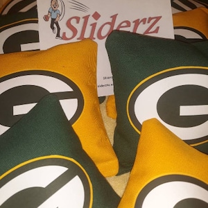 May include: A set of green and gold bean bags with a white "G" logo. The bean bags are labeled "Sliderz" and are perfect for playing cornhole or other games.