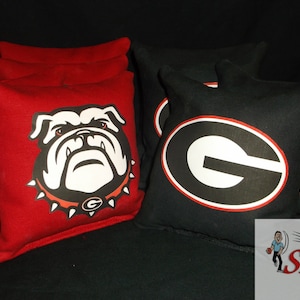 May include: A set of cornhole bags in red and black. The red bags feature a bulldog graphic, while the black bags display a white and red "G" logo. The bags are likely for a sports game.