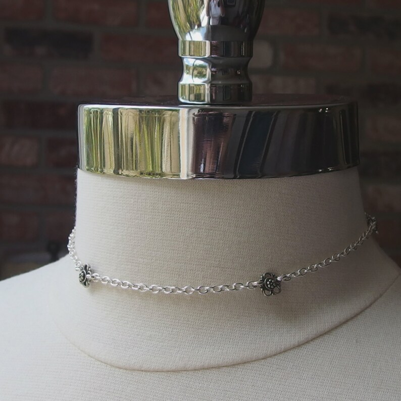 May include: A silver choker necklace with a delicate chain and three small flower charms. The necklace is displayed on a white mannequin bust against a brick background. The flowers have a dark center and detailed petals.