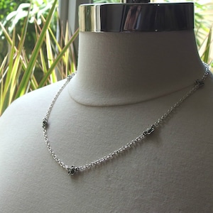 May include: A delicate silver necklace with small, detailed flower charms evenly spaced along the chain. The necklace is displayed on a mannequin, showcasing its design and length. The chain is a classic link style, and the flowers have a dark, contrasting center.