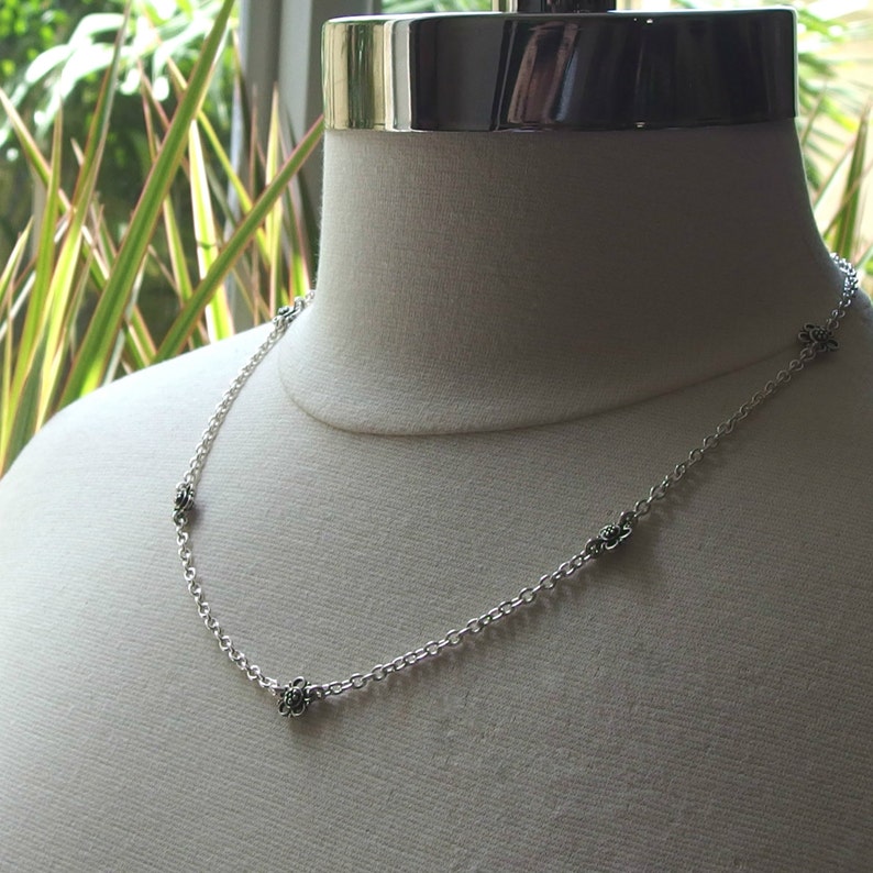 May include: A delicate silver necklace with small, detailed flower charms evenly spaced along the chain. The necklace is displayed on a mannequin, showcasing its design and length. The chain is a classic link style, and the flowers have a dark, contrasting center.