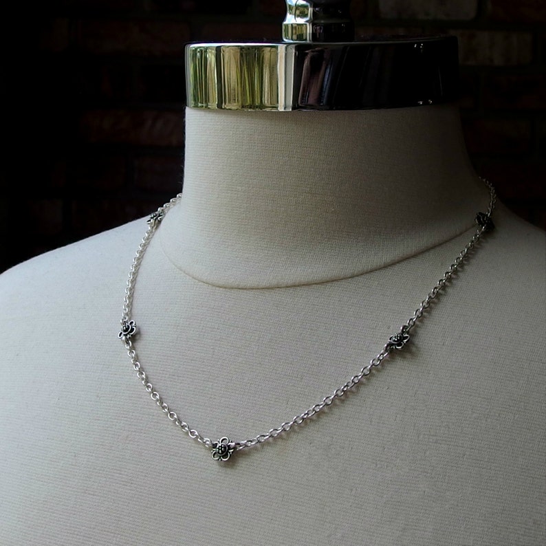 May include: A delicate silver necklace with small, dark floral accents. The chain is fine and the flowers are evenly spaced along the necklace. The necklace is displayed on a white mannequin.