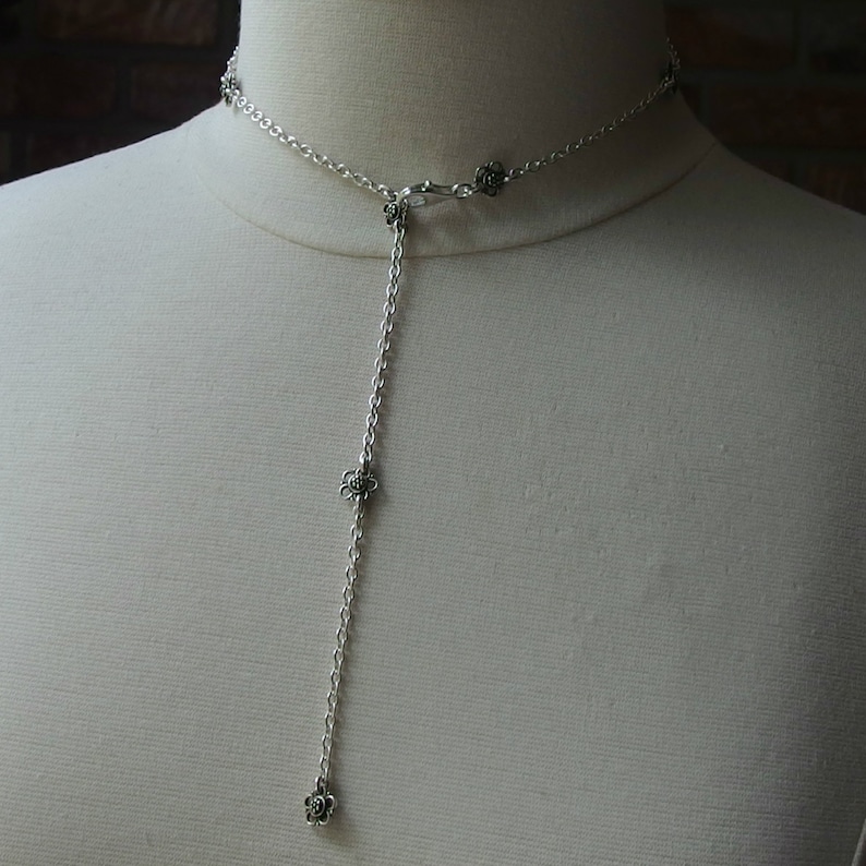 May include: A silver-colored necklace with a delicate chain and a floral pendant design. The necklace features a Y-shaped drop with three flower charms. The necklace is displayed on a mannequin.