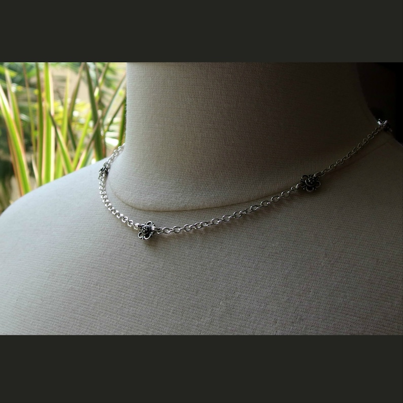 May include: A delicate silver necklace with small flower pendants. The necklace is displayed on a mannequin, showcasing its design. The flowers have dark centers and are evenly spaced along the chain. The background is blurred, with green foliage visible.