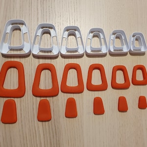 May include: A set of 10 white plastic cookie cutters in various sizes and shapes, along with 10 orange fondant cutouts in the same shapes. The cutters are in the shape of a rounded rectangle with a rounded top and bottom.