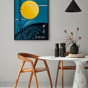 May include: A black framed poster with Japanese text and a yellow moon against a blue sky. The poster is hanging on a white wall above a white marble table with two wooden chairs. There are two black vases with dried flowers on the table.