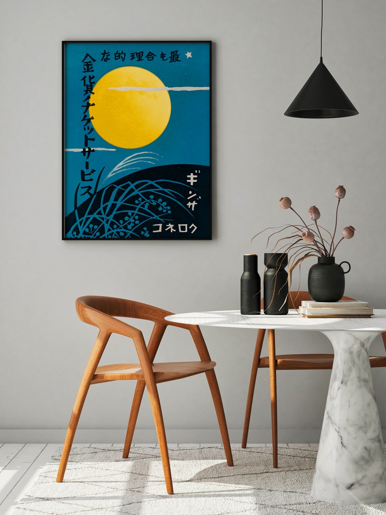 May include: A black framed poster with Japanese text and a yellow moon against a blue sky. The poster is hanging on a white wall above a white marble table with two wooden chairs. There are two black vases with dried flowers on the table.