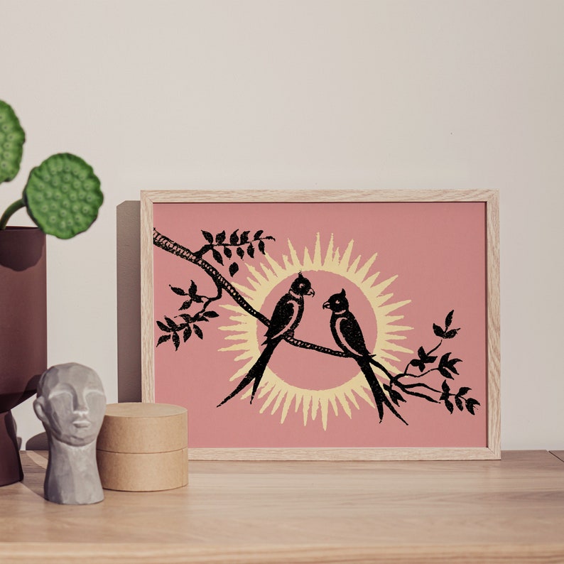 K&ouml;nnte beinhalten: Ein gerahmter Druck mit rosa Hintergrund, auf dem zwei schwarze V&ouml;gel auf einem Ast sitzen, hinter dem sich eine gelbe Sonne befindet.