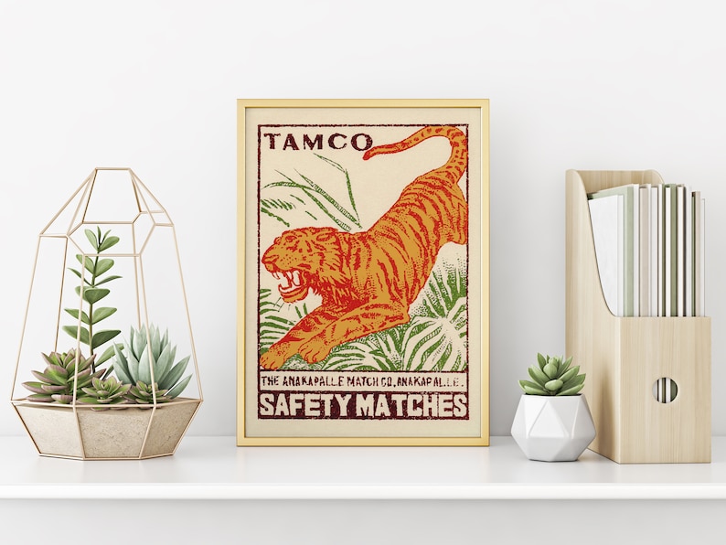 May include: A vintage-style matchbox label featuring a red and orange tiger leaping through green foliage. The label reads "TAMCO THE ANAKAPALLE MATCH CO. ANAKAPALLE. SAFETY MATCHES".