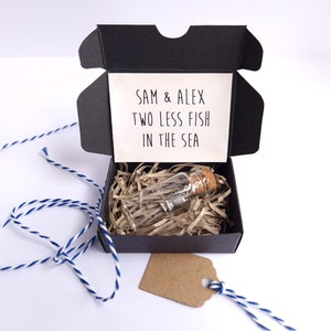 May include: A black gift box with a white card that reads "Sam & Alex Two Less Fish In The Sea". Inside the box is a small glass bottle with a cork stopper and a silver charm.