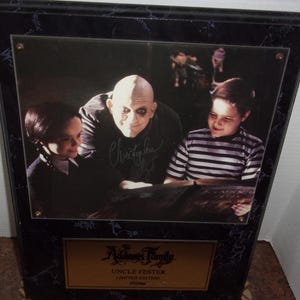 May include: A framed photograph featuring Uncle Fester from The Addams Family, signed by Christopher Lloyd. The image shows Uncle Fester with two children. The frame is black with a marble-like pattern. A gold plaque at the bottom reads "Addams Family" and "Uncle Fester Limited Edition".