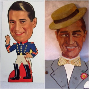 May include: Two vintage cardboard cutouts of a smiling man. One is dressed in a blue military uniform with gold buttons and white pants. The other wears a gray suit, a yellow hat, and a polka-dot bow tie. Both have a cartoonish style.