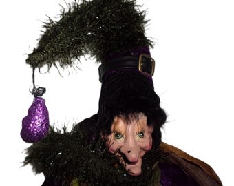 Könnte beinhalten: Eine skurrile Hexenfigur mit einem schwarzen Hut, verziert mit einer Schnalle und einem flauschigen Besatz. Die Hexe trägt einen lila Samtumhang und hat einen grünen und braunen Kragen. Ein lila glitzernder Beutel hängt am Hut.