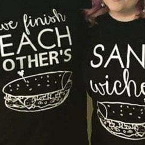 Op de afbeelding: Twee zwarte t-shirts met witte tekst en sandwich illustraties. E&eacute;n shirt leest "we finish EACH OTHER'S" boven een sandwich afbeelding. Het andere shirt zegt "SANDwiches" met een bijpassende sandwich afbeelding.