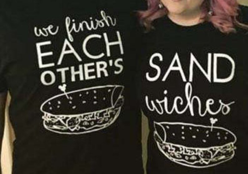Op de afbeelding: Twee zwarte t-shirts met witte tekst en sandwich illustraties. E&eacute;n shirt leest "we finish EACH OTHER'S" boven een sandwich afbeelding. Het andere shirt zegt "SANDwiches" met een bijpassende sandwich afbeelding.