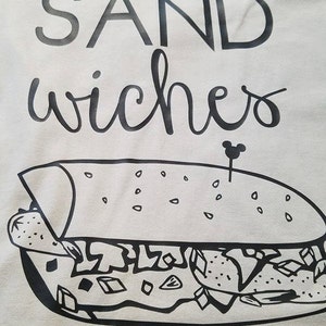 Op de afbeelding: Lichtgrijs t-shirt met het woord "SANDwiches" in zwarte, gestileerde letters. Daaronder een zwart-wit illustratie van een sandwich met een Mickey Mouse-vormige tandenstoker.
