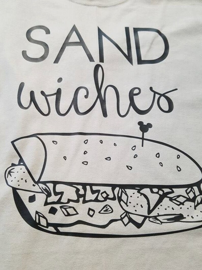 Op de afbeelding: Lichtgrijs t-shirt met het woord "SANDwiches" in zwarte, gestileerde letters. Daaronder een zwart-wit illustratie van een sandwich met een Mickey Mouse-vormige tandenstoker.