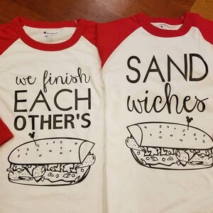 Op de afbeelding: Twee witte en rode raglan shirts met zwarte tekst en sandwich illustraties. E&eacute;n shirt zegt "we finish EACH OTHER'S" en de andere zegt "SAND wiches". De shirts hebben een Champion label.