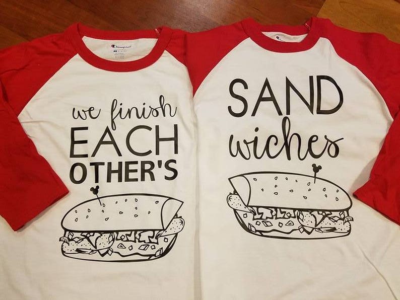 Op de afbeelding: Twee witte en rode raglan shirts met zwarte tekst en sandwich illustraties. E&eacute;n shirt zegt "we finish EACH OTHER'S" en de andere zegt "SAND wiches". De shirts hebben een Champion label.