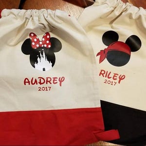 May include: Two drawstring bags with red bottoms and straps. One bag features a Minnie Mouse design with the name "Audrey" and the year "2017". The other bag has a pirate Mickey Mouse design with the name "Riley" and the year "2017".