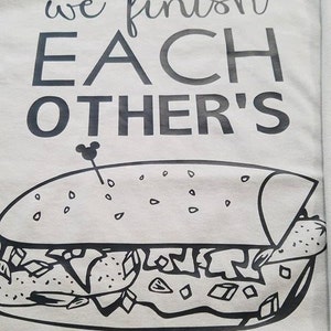 Op de afbeelding: Lichtgrijs t-shirt met de tekst "we finish EACH OTHER'S" boven een zwart-witte afbeelding van een sandwich. Een klein Mickey Mouse-hoofd staat naast het woord "OTHER'S".