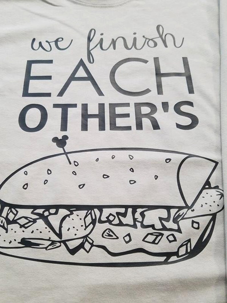 Op de afbeelding: Lichtgrijs t-shirt met de tekst "we finish EACH OTHER'S" boven een zwart-witte afbeelding van een sandwich. Een klein Mickey Mouse-hoofd staat naast het woord "OTHER'S".