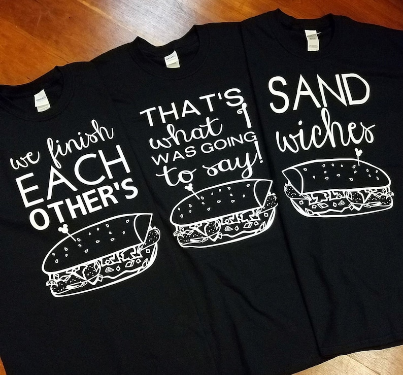 Op de afbeelding: Drie zwarte t-shirts met witte tekst en sandwich illustraties. E&eacute;n shirt leest "we finish EACH OTHER'S", een ander zegt "THAT'S what I WAS GOING to say!", en de derde zegt "SAND wiches".