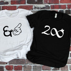 Puede incluir: Dos camisetas, una blanca y otra negra, se muestran contra una pared de ladrillos. La camiseta blanca presenta una & negra y una mano de dibujos animados. La camiseta negra tiene el n&uacute;mero 2 y un s&iacute;mbolo de infinito en blanco.