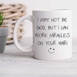 Pode incluir: Caneca de café de cerâmica branca com texto preto que diz "Eu posso não ser Deus, mas posso fazer milagres no seu cabelo!" e um rosto sorridente.