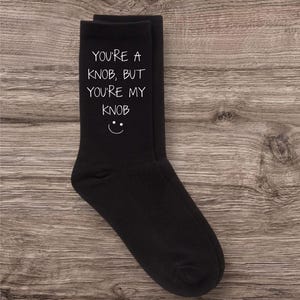 May include: A pair of black socks featuring the white text "YOU'RE A KNOB, BUT YOU'RE MY KNOB" and a smiley face. The socks are presented on a wooden background.