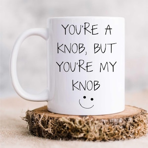 May include: White ceramic coffee mug with black text that reads "You're a knob, but you're my knob" and a simple smiley face.