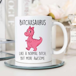 May include: White ceramic mug with the text "BITCHASAURUS" above a pink cartoon dinosaur wearing a bow. Below the dinosaur is the text "LIKE A NORMAL BITCH, BUT MORE AWESOME". The mug has a white handle and sits on a clear, round coaster.
