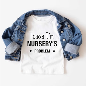 May include: A white t-shirt with the text "Today I'm NURSERY'S PROBLEM" in black, with stars above and below the word "PROBLEM". The shirt is paired with a blue denim jacket.