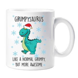 Op de afbeelding: Witte keramische mok met een cartoon dinosaurus met een kerstmuts. De mok heeft de tekst "GRUMPYSAURUS" boven de dinosaurus en "LIKE A NORMAL GRUMPY, BUT MORE AWESOME" eronder. Blauwe sneeuwvlokken versieren de mok.