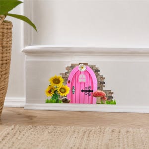 May include: A whimsical pink fairy door with a white daisy on top, set into a brick wall. Sunflowers, a ladybug, a snail, and red-capped mushrooms decorate the scene. The door has a small window and black hardware.