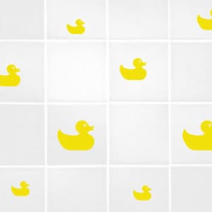 Op de afbeelding: Een witte betegelde badkamermuur met gele rubberen eendenstickers. De eenden zijn in verschillende maten en posities.