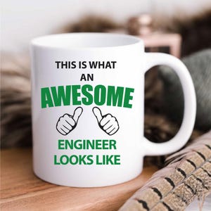 May include: White ceramic coffee mug with a green and black design. The mug reads "This is what an awesome engineer looks like" with two thumbs up symbols.