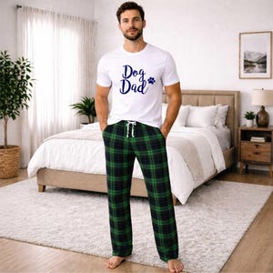 May include: White t-shirt with "Dog Dad" text and paw print graphic. Paired with green and black plaid pajama pants. The pants have a drawstring waist. The outfit is casual and comfortable.