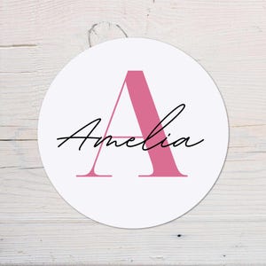 Peut inclure: Un dessous de verre circulaire blanc avec la lettre "A" rose et le nom "Amelia" en écriture noire. Le design est centré sur le dessous de verre, sur un fond clair en bois.