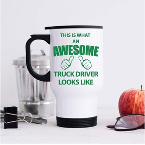 Könnte beinhalten: Weißer Reisebecher mit schwarzem Griff, Deckel und Boden. Auf dem Becher steht in grüner Schrift "THIS IS WHAT AN AWESOME TRUCK DRIVER LOOKS LIKE". Der Becher steht auf einer weißen Oberfläche.
