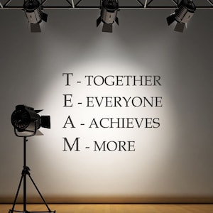 May include: A white wall with the acronym TEAM spelled out with the words "Together", "Everyone", "Achieves", and "More" written below each letter. The wall is lit by spotlights.
