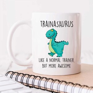 May include: White ceramic coffee mug featuring a green cartoon dinosaur and the text "TRAINASAURUS LIKE A NORMAL TRAINER, BUT MORE AWESOME".