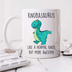 May include: White ceramic mug featuring a teal dinosaur illustration with a green belly. The mug has the text "KNOBASAURUS" and "LIKE A NORMAL KNOB, BUT MORE AWESOME". The dinosaur is smiling. A fun, novelty item.