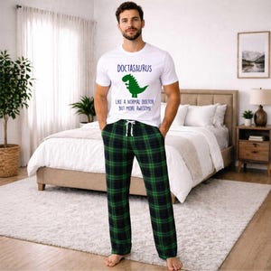 May include: White t-shirt with the text "DOCTASAURUS" and a green dinosaur graphic. The shirt is paired with green and black plaid pajama pants. The pajama pants have a drawstring waist.