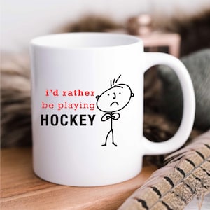 May include: White ceramic coffee mug with a black and white illustration of a stick figure with a sad face. The text "i'd rather be playing HOCKEY" is printed in black and red on the mug.