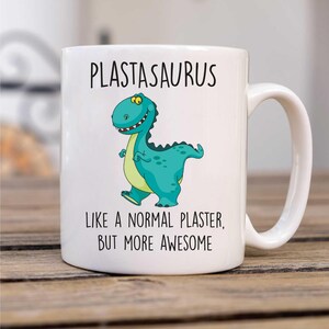 May include: White ceramic mug with a green cartoon dinosaur and the text "PLASTASAURUS LIKE A NORMAL PLASTER, BUT MORE AWESOME".