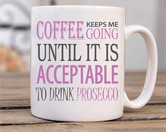May include: White ceramic coffee mug with the text "Coffee keeps me going until it is acceptable to drink Prosecco" in black and pink lettering.