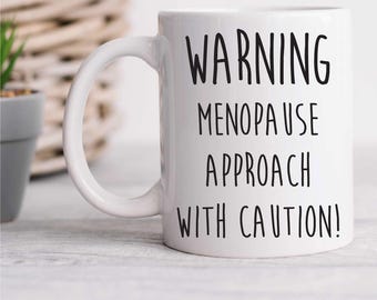 May include: White ceramic coffee mug with black text that reads "WARNING MENOPAUSE APPROACH WITH CAUTION!"
